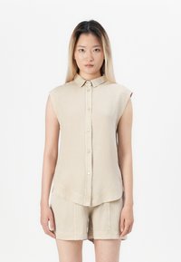Camicia senza maniche color beige, tessuto morbido, colletto semplice, orlo curvo, tasche frontali a toppa, abbinata a pantaloni corti coordinati.