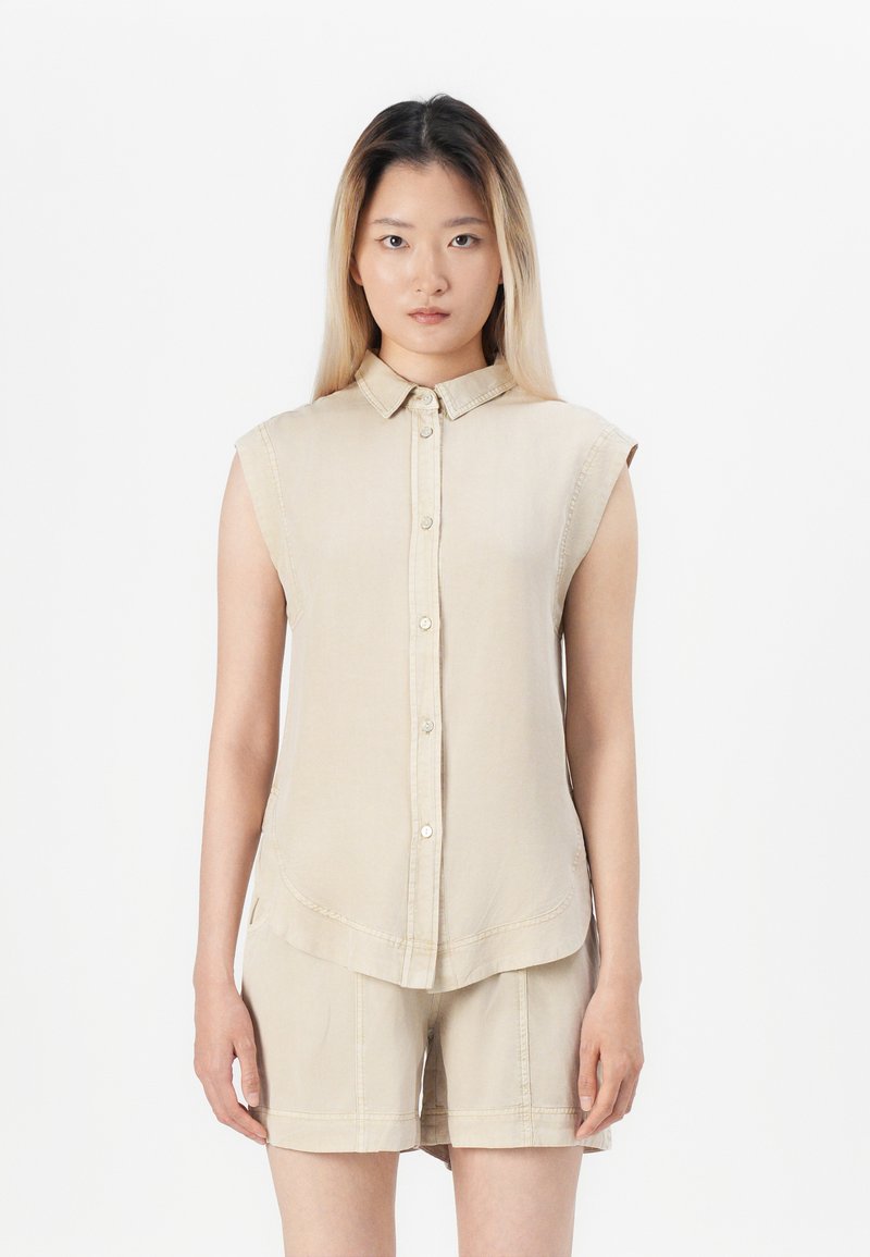 Camicia senza maniche color beige, tessuto morbido, colletto semplice, orlo curvo, tasche frontali a toppa, abbinata a pantaloni corti coordinati.