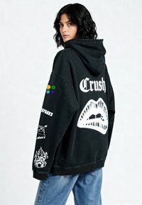 Crna prevelika dukserica s kapuljačom s natpisom "Crush" u bijeloj boji, koja sadrži grafiku usana, šarene zakrpe i razne printane detalje na rukavima. Tkanina od mješavine pamuka.