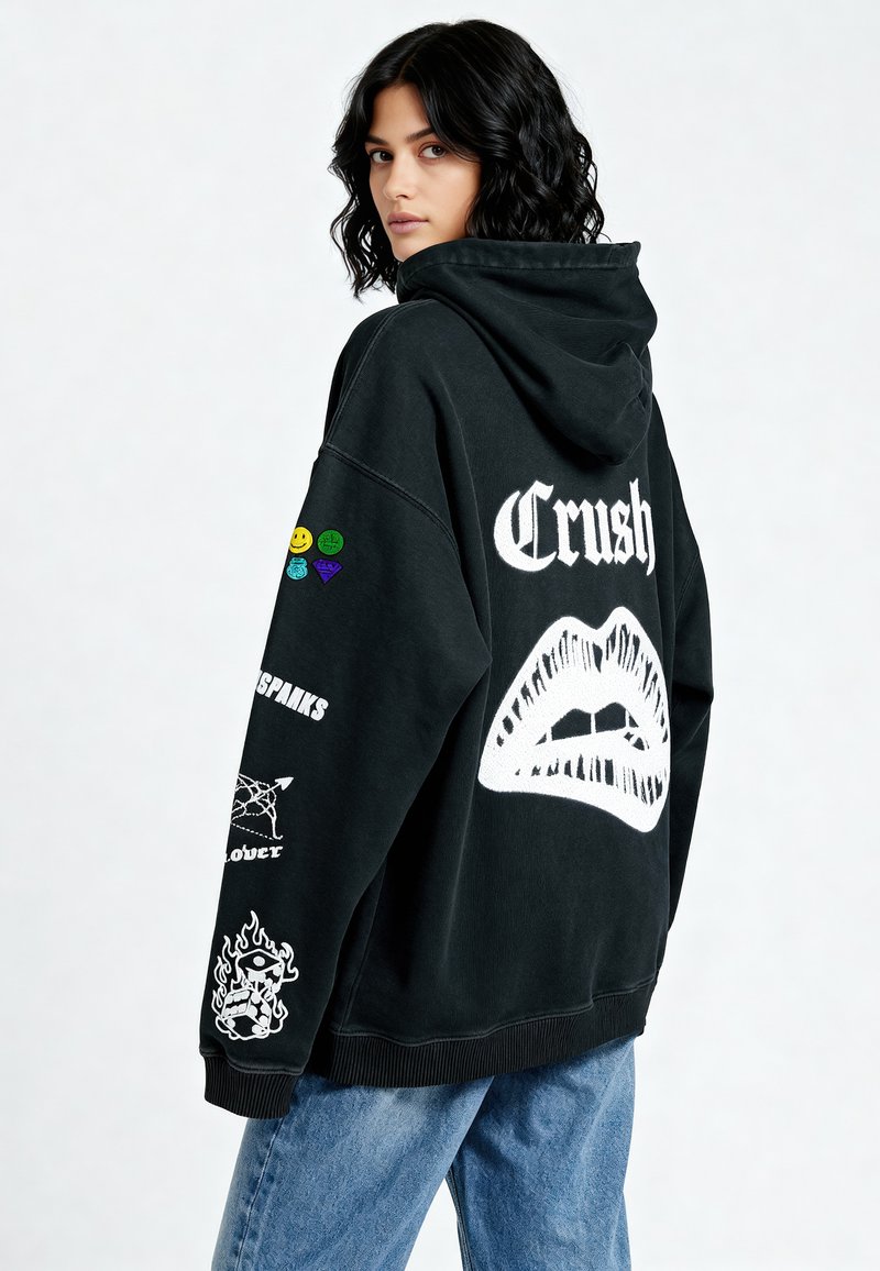 Crna prevelika dukserica s kapuljačom s natpisom "Crush" u bijeloj boji, koja sadrži grafiku usana, šarene zakrpe i razne printane detalje na rukavima. Tkanina od mješavine pamuka.