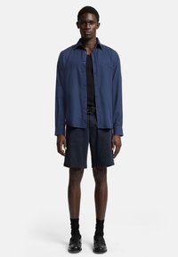 Chemise boutonnée bleu marine à manches longues, portée sur un débardeur noir. Associée à un short sombre et des chaussures noires avec des chaussettes.
