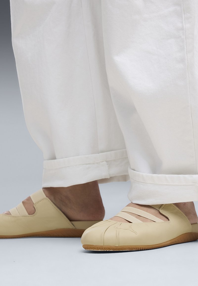 Πόδια που φορούν μπεζ παπούτσια τύπου slip-on με ελαστικές λουρίδες και στρογγυλεμένες μύτες, σε συνδυασμό με λευκό, φαρδύ παντελόνι με γυρισμένες μανσέτες.