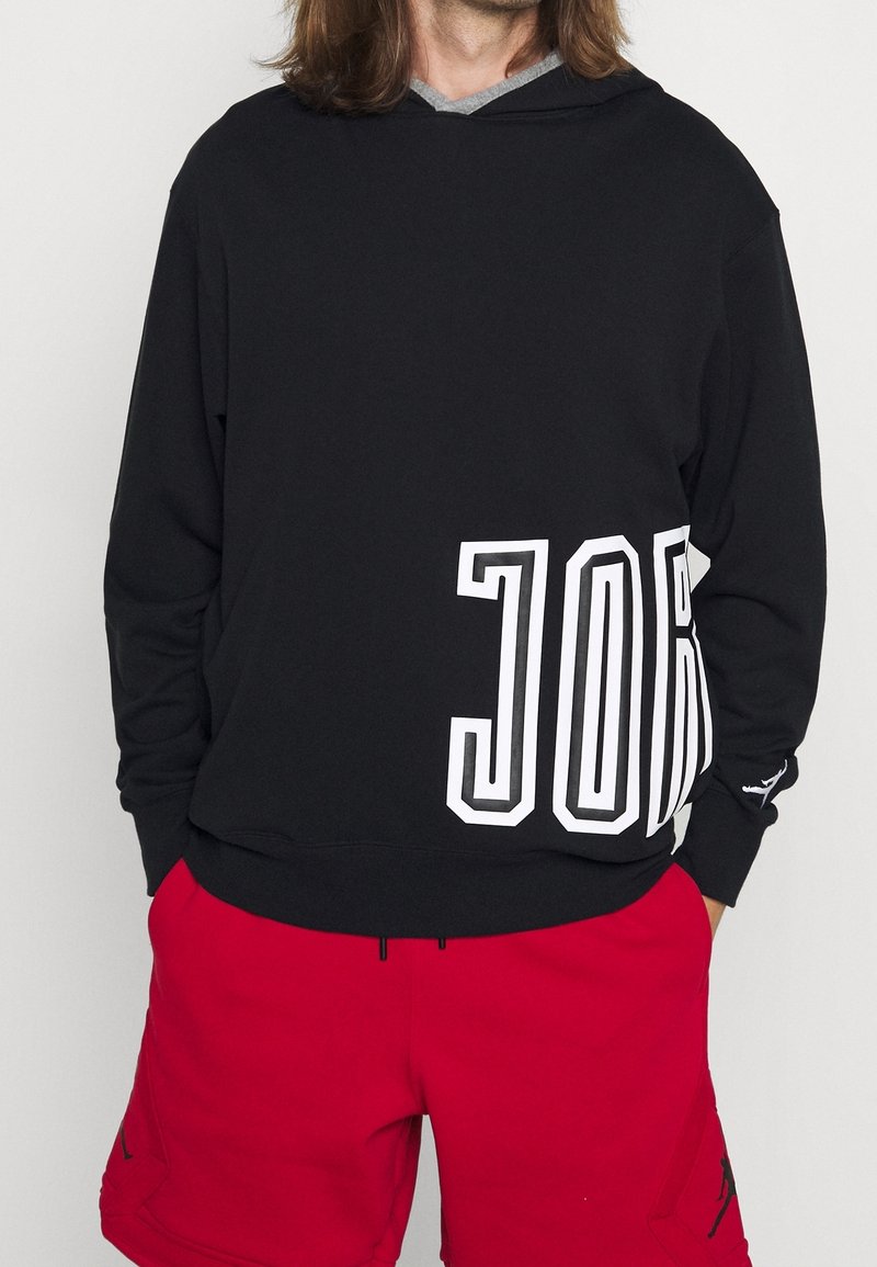 Personne portant un sweat à capuche noir avec un grand texte blanc « JOR » sur le côté et un short rouge avec des poches latérales.