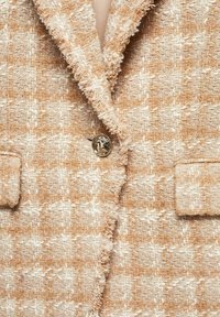 Veste en tweed beige et blanc texturée avec des revers effilochés, un bouton décoratif unique et deux poches à rabat.