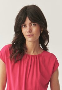 Rosa bluse med korte ærmer, løs pasform og samlet halsdetalje. Modellen bærer en guld geometrisk ørering. Baggrunden er ensfarvet hvid.