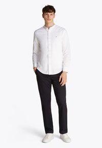 Camicia bianca con bottoni e colletto alla coreana, abbinata a pantaloni blu navy e sneakers bianche. La camicia presenta un piccolo logo sul petto.