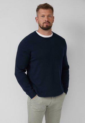 Man met een baard, gekleed in een marineblauwe trui over een wit shirt en beige broek, staande met handen in zijn zakken tegen een effen achtergrond.
