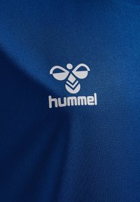 Blått tyg med en slät textur har en vit Hummel-logotyp, som består av en stiliserad bi och varumärkesnamnet.