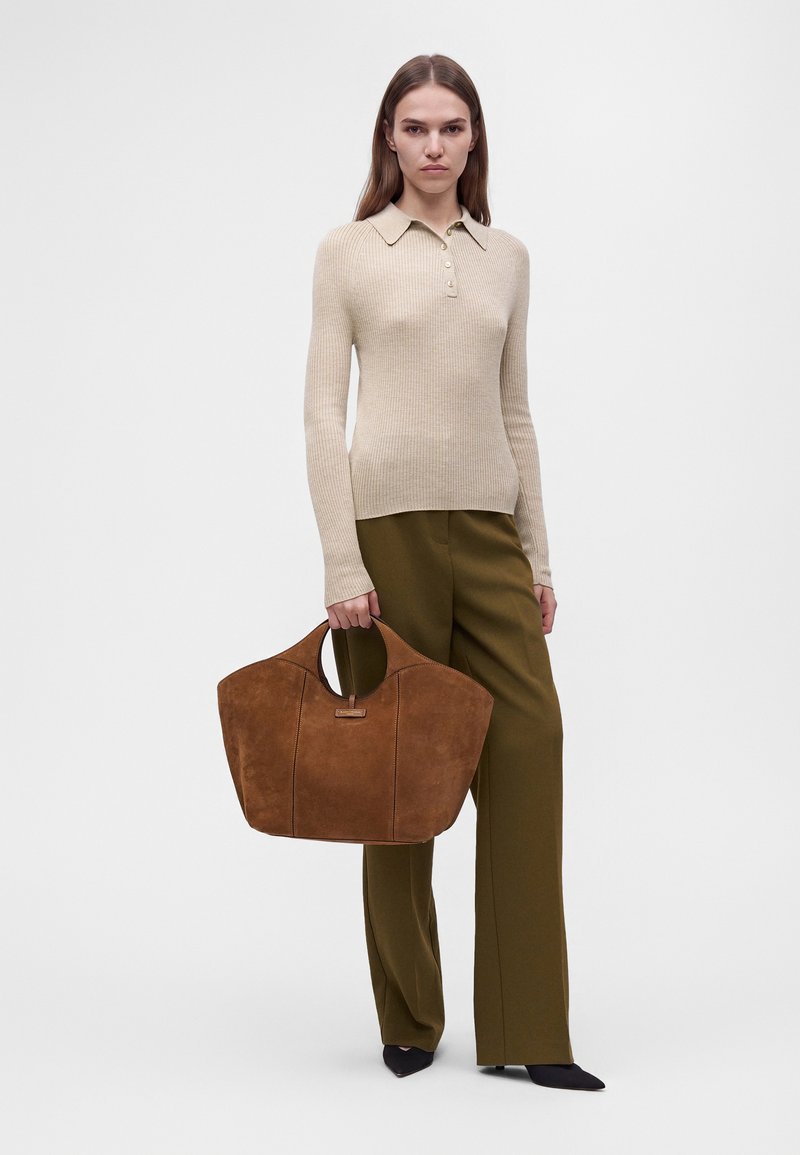 Femme portant un haut beige à manches longues côtelé et un pantalon olive, tenant un grand sac cabas en daim marron avec des poignées rondes.