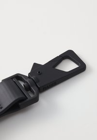 Schwarze Kunststoffschnalle mit geometrischem Design. Verfügt über einen schmalen Riemen und einen auffälligen Befestigungsmechanismus. Die Marke "RAINS" ist eingeprägt.