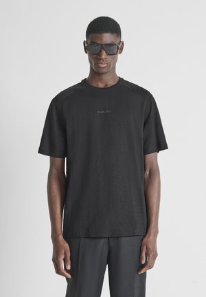 Tall person wearing black oversized sunglasses, black short-sleeve shirt with "MORATO" text, and black pants standing against plain background.