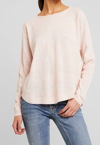 Hellrosa gestrickter Pullover mit lockerem Sitz. Verfügt über strukturierte horizontale Streifen und lange Ärmel, kombiniert mit blauen Jeans.