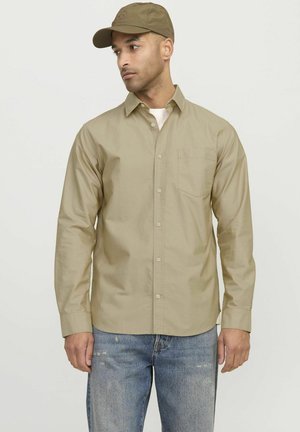 Camisa de botón color caqui claro con mangas largas, un solo bolsillo en el pecho, combinada con una gorra a juego. Llevada con jeans azules deslavados.
