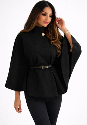 Femme portant un manteau cape noir ceinturé avec de larges manches et un bouton doré près du col, assorti à un pantalon noir.