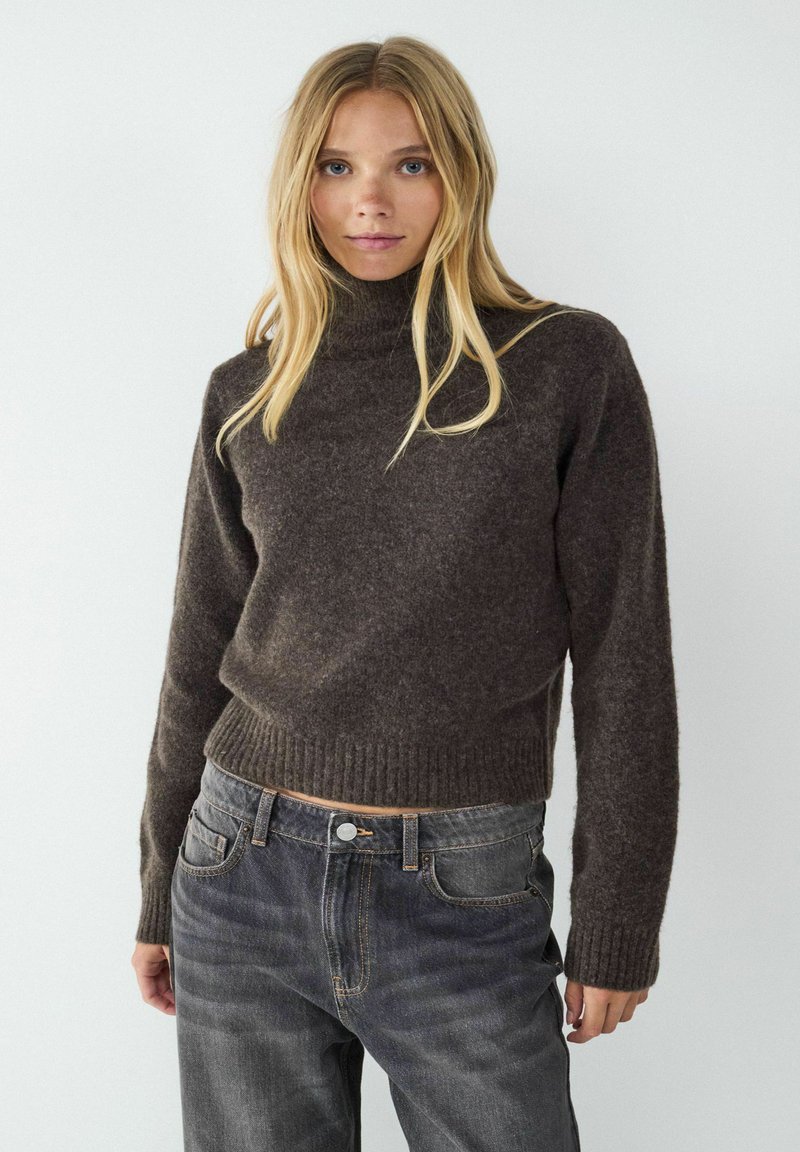 Brauner Rollkragenpullover mit geripptem Saum, aus weichem Material, kombiniert mit dunklen Jeans, die Taschen und einen Knopfverschluss haben.