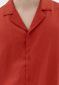 Kragengesegneter roter Hemdblusenshirt mit drei sichtbaren Knöpfen und einem offenen Ausschnitt, der teilweise die Brust zeigt.