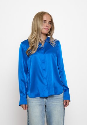 Giovane donna con lunghi capelli biondi che indossa una camicetta di raso blu brillante e jeans azzurri chiari su uno sfondo bianco semplice.