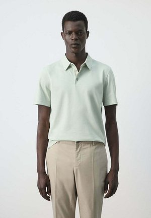 Hombre de pie con una camisa polo de manga corta verde claro y pantalones beige frente a un fondo blanco liso.