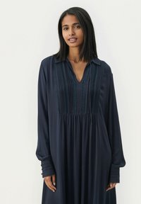 Marineblaues Langarmkleid mit V-Ausschnitt, ausgestattet mit vertikalen Spitzendetails und einer gerafften Taille. Die Manschettenakzente mit Knöpfen verleihen dem Kleid zusätzliche Details.
