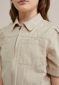 Bežna zip-up majica s kratkimi rokavi, ki ima dva žepa na prsih, usnjeno ovratnico in subtilne šivane detajle.