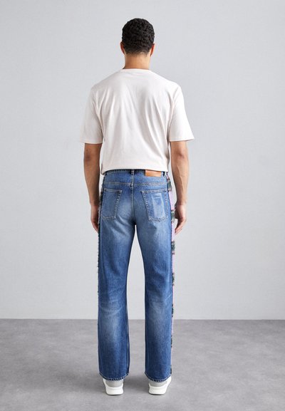 Jean en denim bleu à coupe droite, avec des accents en tissu multicolore sur les côtés et une étiquette en cuir marron à l'arrière de la taille.