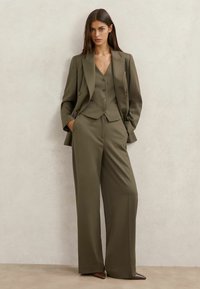 Una donna è in piedi contro una parete chiara, indossando un completo verde oliva con blazer doppiopetto, gilet abbottonato, pantaloni a gamba larga e scarpe marroni con punta appuntita.