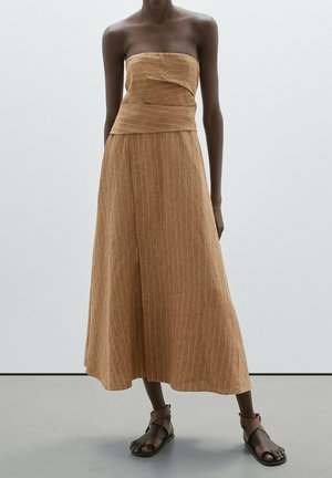 Robe marron sans bretelles, à fines rayures, longueur cheville, avec un corsage drapé asymétrique portée avec des sandales marron à bride à la cheville sur une personne à la peau foncée.