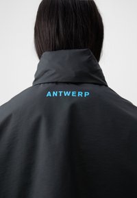 Black jacket with a high collar, featuring the text "ANTWERP" in bright blue on the back. Smooth texture and minimalist design.