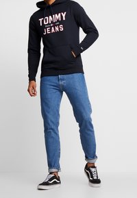 Muž v černé mikině Tommy Jeans, modrých zúžených džínách a černo-bílých teniskách stojící proti jednobarevnému světlému pozadí.