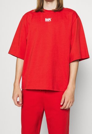 Personne portant un T-shirt rouge ample avec le logo "MA Martin Asbjurn" et un pantalon rouge assorti sur un fond clair uni.