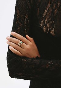 Anillo de oro con una piedra de esmeralda, que presenta superficies texturizadas y lisas, llevado en una mano que descansa sobre una manga de encaje negro.