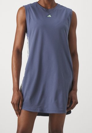 Granatowa sportowa sukienka bez rękawów z białymi paskami po bokach, noszona przez osobę stojącą na tle jednolitego jasnego tła.