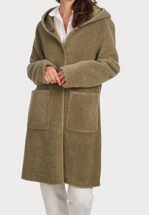 Femme portant un manteau duveteux vert olive longueur genou avec de grandes poches avant sur une chemise blanche et un pantalon clair.