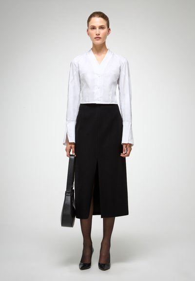 Blusa bianca a maniche lunghe, corta, con scollo a V e chiusura a bottoni, abbinata a una gonna nera aderente al ginocchio con uno spacchetto frontale. Borsa nera e tacchi.