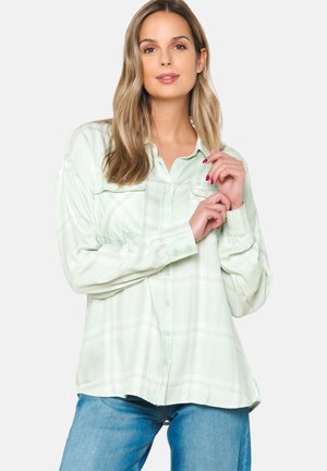 Mint green plaid button-up shirt with two chest pockets, soft fabric, long sleeves, and a curved hemline, paired with blue jeans.