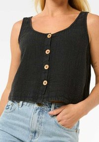 Nainen, jolla on yllään hihaton musta napitettu crop-top ja vaaleansiniset korkeavyötäröiset farkut, yhdellä kädellään taskussa.