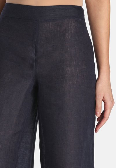 Vrouw die hoge taille, donkerblauwe linnenbroek met rechte pasvorm draagt, waarbij de taille tot halverwege de dij zichtbaar is en de rechterhand ontspannen langs het lichaam hangt.