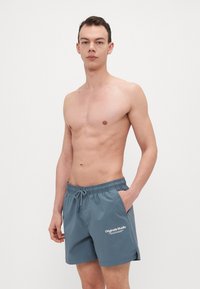 Costumi da nuoto da uomo in tessuto teal, dotati di vita elasticizzata con cordoncino e tasche laterali. Accentuati da un dettaglio del logo bianco.