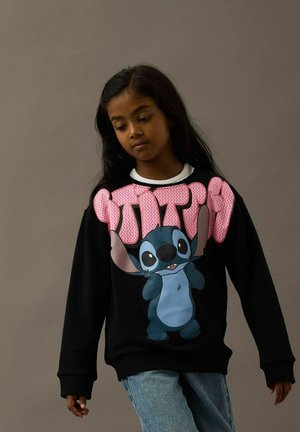 Ung pige med langt, mørkt hår iført en sort sweatshirt med store lyserøde bogstaver og en blå tegneseriefigur, parret med blå jeans.