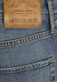 Jeans de mezclilla azul con un parche de cuero marrón claro que presenta texto en relieve. La costura distintiva y las zonas desgastadas realzan la textura de la tela.