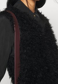 Gilet in pelliccia sintetica nera con una finitura testurizzata, abbinato a una camicia nera. Presenta una cinghia bordeaux sul lato sinistro per un dettaglio in più.