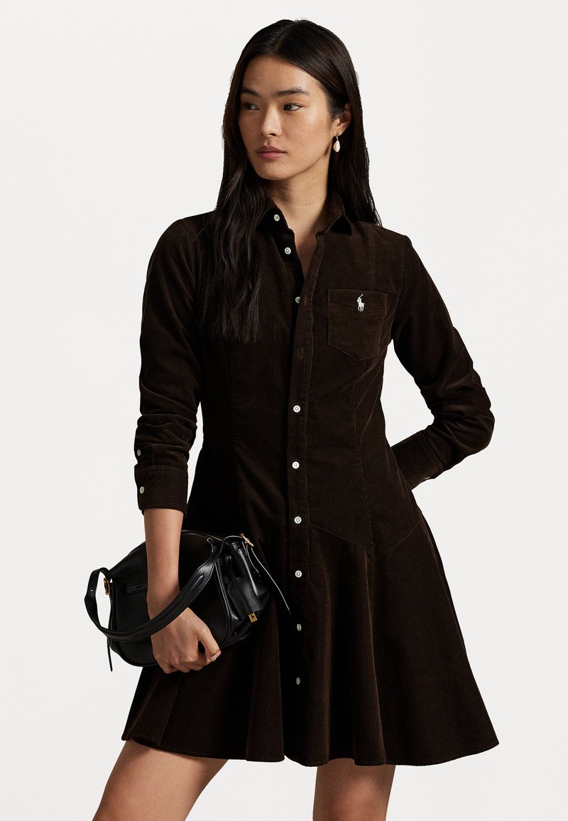 Brown corduroy shirt dress with long sleeves, button-down front, and a chest pocket. Accented with a small logo and flared hem.