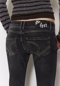 Črne kavbojke iz jeansa s srebrnimi žebljiči na žepih, standardnim pasom in etiketom z napisom "sixteen." Tkanina je videti rahlo obrabljena.