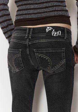 Črne kavbojke iz jeansa s srebrnimi žebljiči na žepih, standardnim pasom in etiketom z napisom "sixteen." Tkanina je videti rahlo obrabljena.