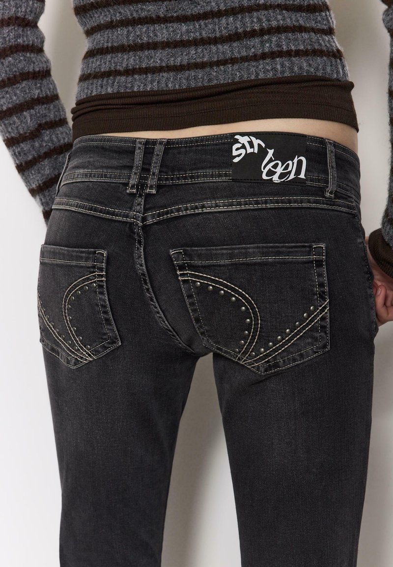 Črne kavbojke iz jeansa s srebrnimi žebljiči na žepih, standardnim pasom in etiketom z napisom "sixteen." Tkanina je videti rahlo obrabljena.