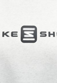 Biely textil s jemnou textúrou, s odvážnym čiernym logom a textom "KE" a "SH" s geometrickými dizajnovými prvkami.