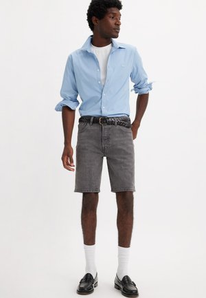 Camicia azzurra chiara a maniche lunghe, t-shirt bianca, pantaloni corti in denim grigio, cintura intrecciata nera, calze bianche e mocassini neri indossati da un modello in piedi.