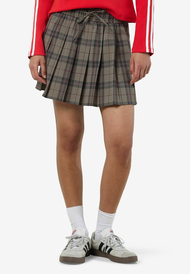 Gonna a pieghe a quadretti grigi con vita regolabile, con accenti rossi e blu. Indossata con calzini bianchi e sneakers grigie.