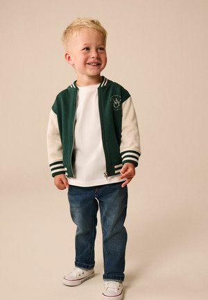 Young blonde boy smiling, wearing green and white varsity jacket, white t-shirt, blue jeans, and white sneakers, standing on a beige background.