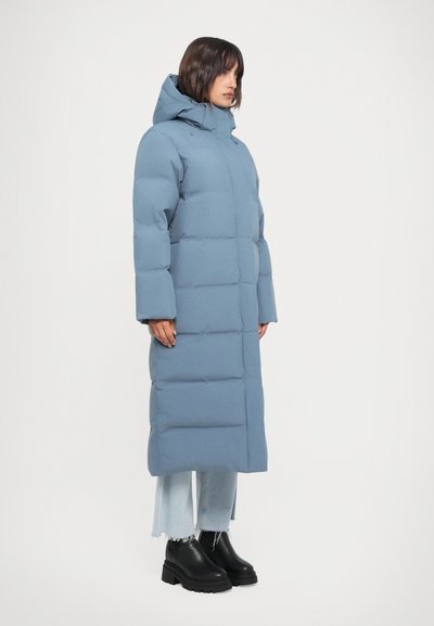 Long, puffy blue coat with a hood, featuring a boxy shape and thick padding; worn over faded jeans and chunky black boots.